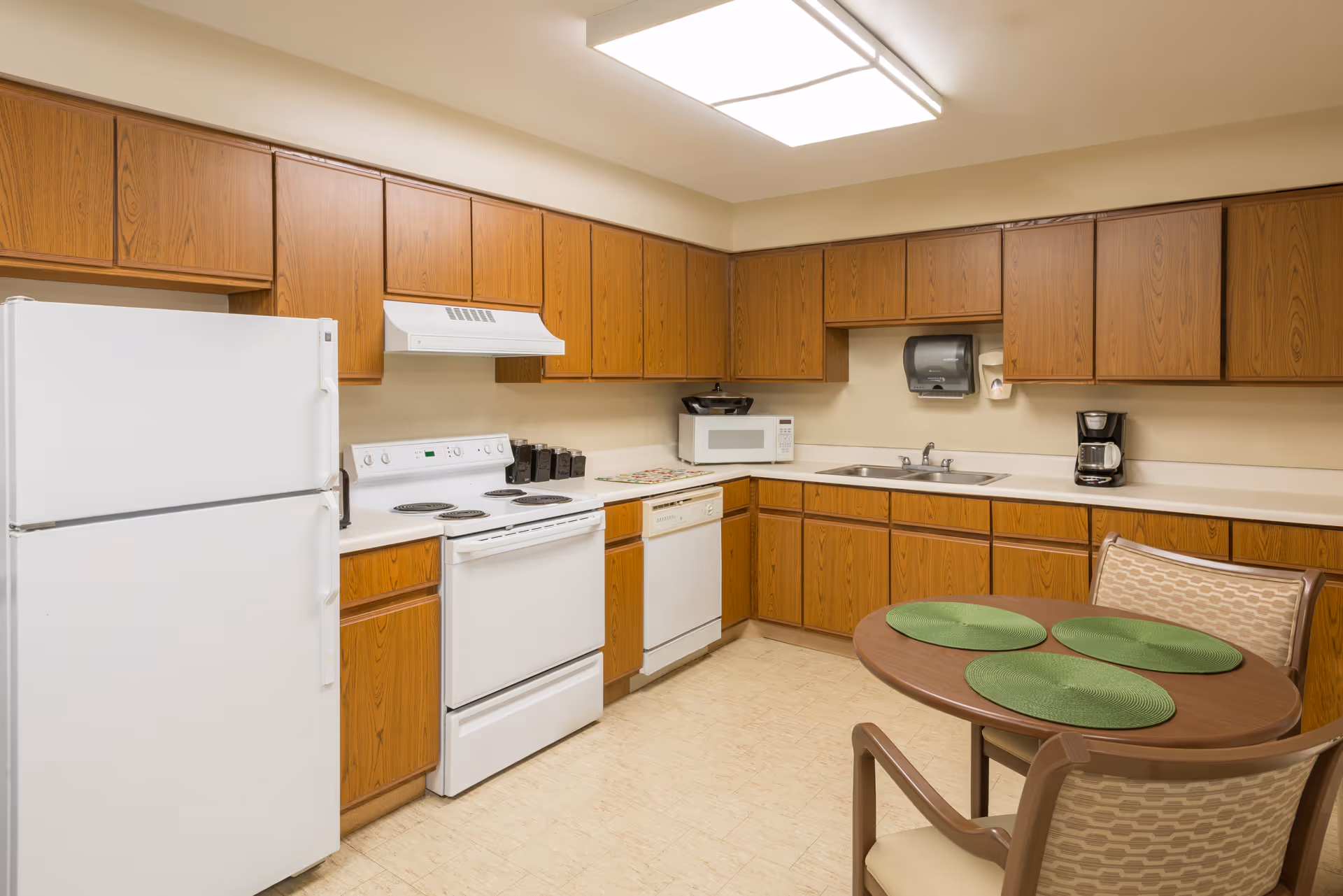 Well-lit institutional kitchen with wooden cabinets, white appliances, a sink and a small round table with green placemats.