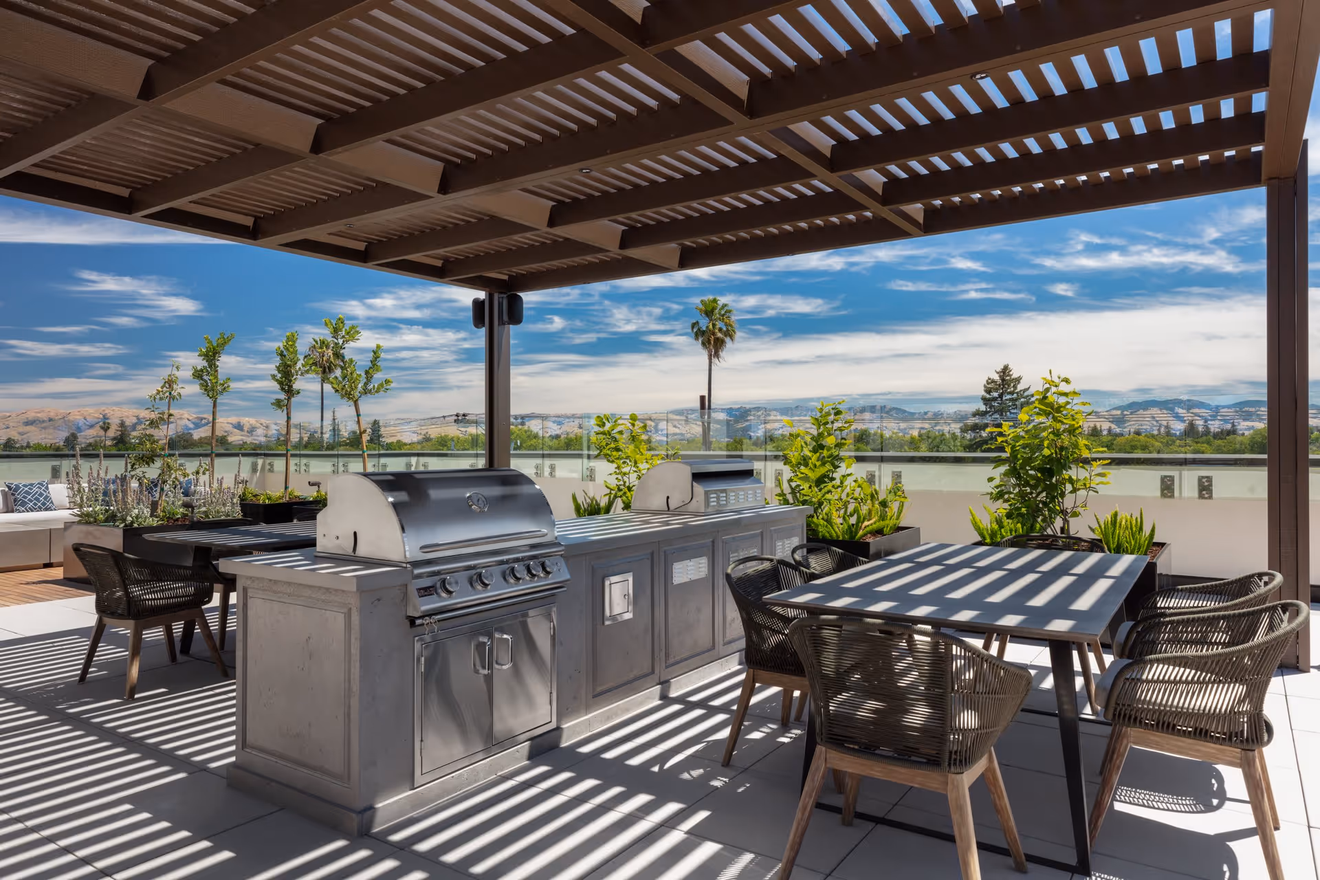 Outdoor patio area with a pergola overhead casting striped shadows, featuring a stainless steel barbecue grill and countertop, a dining table with four woven chairs, potted plants, and a scenic view of distant hills and a partly cloudy sky.