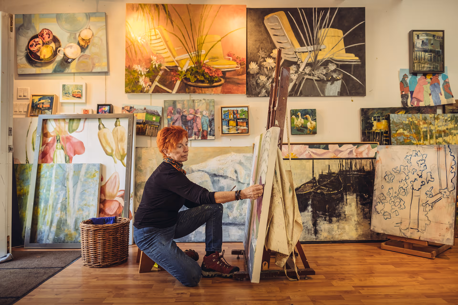 A woman with short red hair kneeling on the floor and painting on a canvas in an art studio filled with various colorful paintings displayed on the walls and floor.