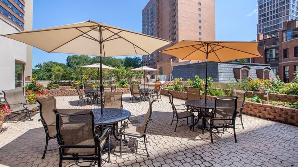 Outdoor patio area with multiple round metal tables and mesh chairs, each table shaded by large beige umbrellas. The patio is paved with stone tiles and surrounded by raised garden beds with greenery and flowers. Tall buildings and a clear blue sky are visible in the background.