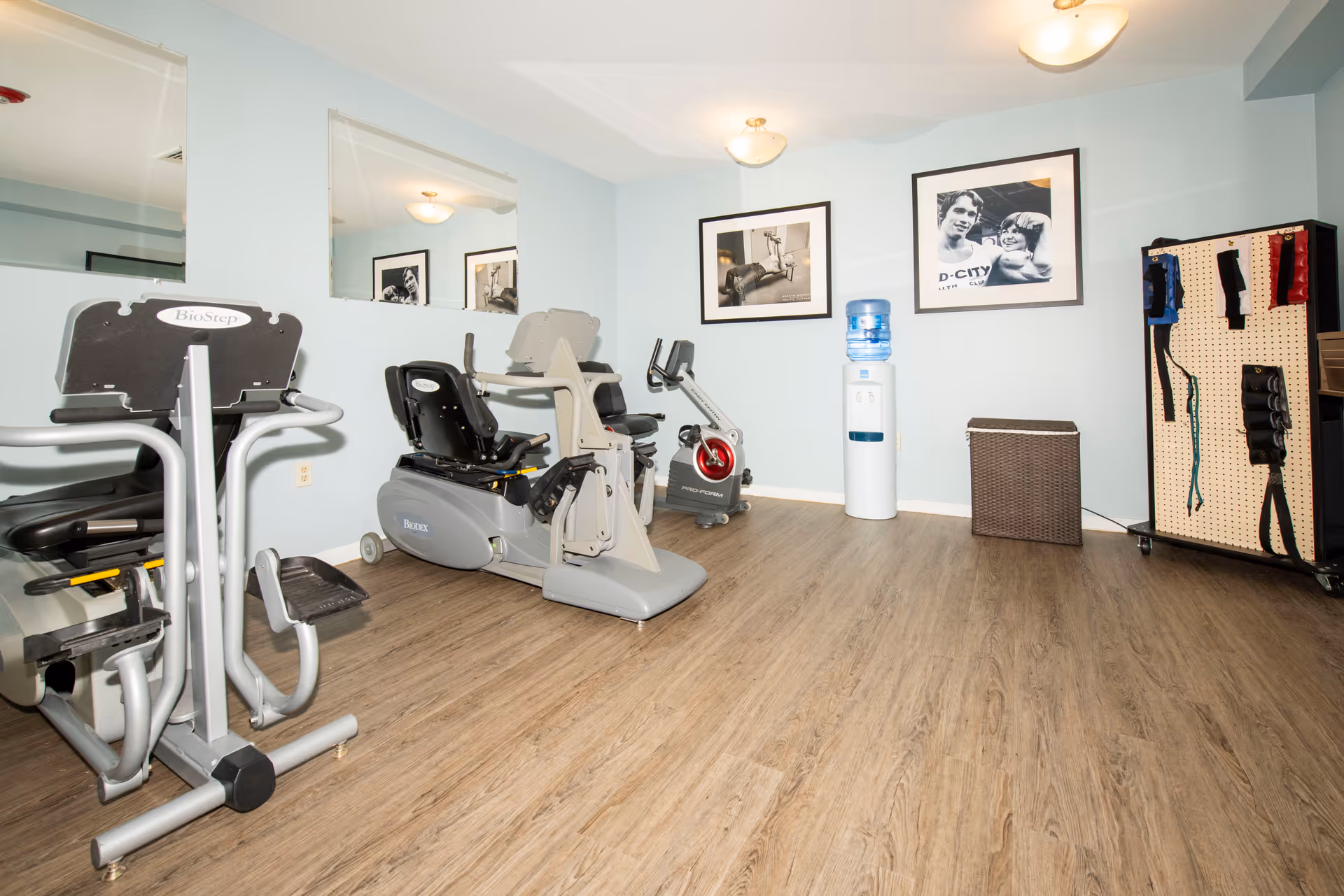 A small exercise room with light blue walls and wood flooring. The room contains several pieces of exercise equipment including a BioStep machine, a recumbent bike, and a stationary bike. There is a water cooler and a wicker basket against the far wall. Two black and white framed photos hang on the wall above the water cooler. On the right side, there is a pegboard with various exercise bands and straps hanging on it. Two ceiling lights illuminate the room.