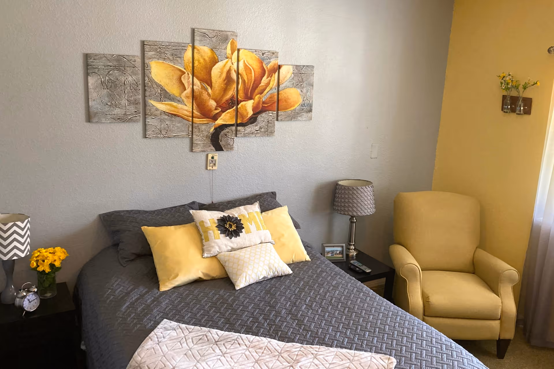 A cozy bedroom with a gray bedspread and yellow accent pillows, including one with the word 'HOME' on it. Above the bed is a multi-panel wall art featuring a large yellow flower. On either side of the bed are nightstands with lamps, a small clock, and a vase with yellow flowers. To the right of the bed is a comfortable yellow armchair next to a window with sheer curtains and a small wall decoration with flowers.