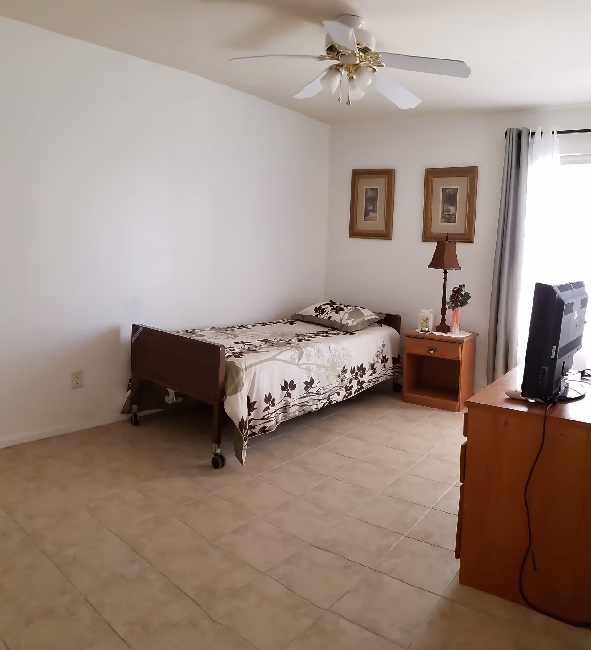 Single-occupancy bedroom with a wheeled bed, nightstand, lamp, and a TV on a dresser.