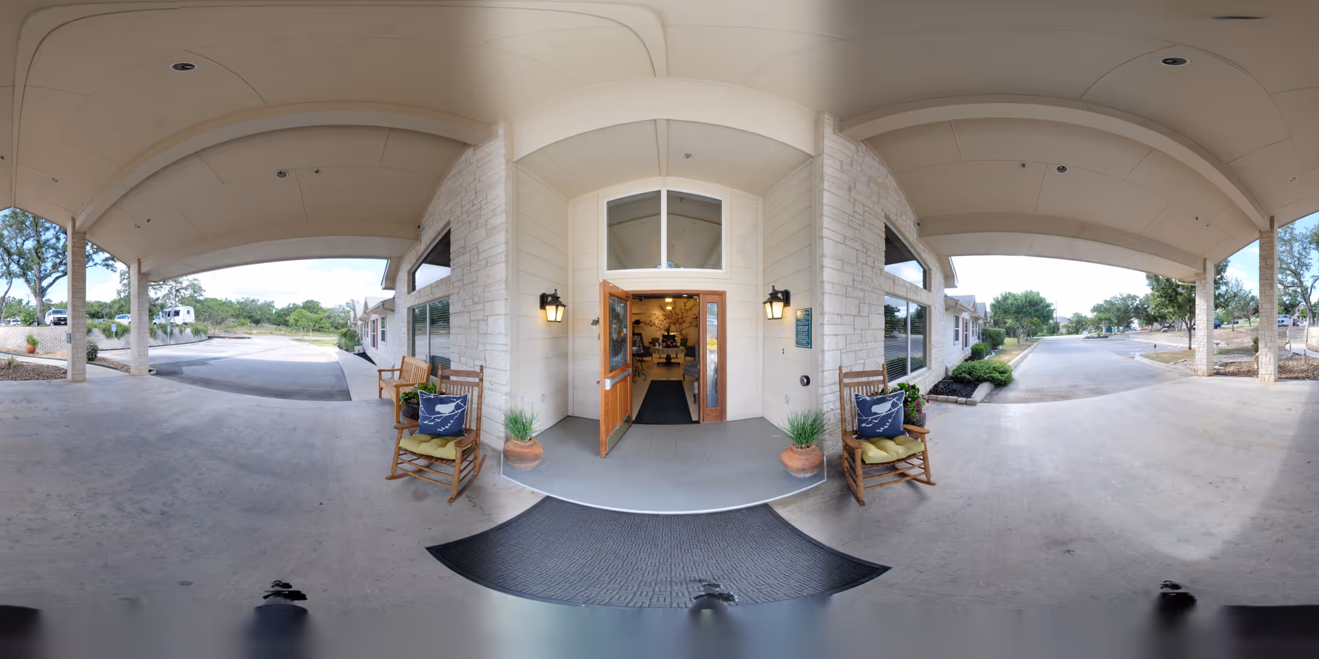Covered entrance area of Heritage Place of Boerne with two wooden rocking chairs on either side of the open double wooden doors. The entrance is flanked by stone and light-colored siding walls, with potted plants and outdoor wall lanterns. The driveway and surrounding greenery are visible in the background under a clear sky.
