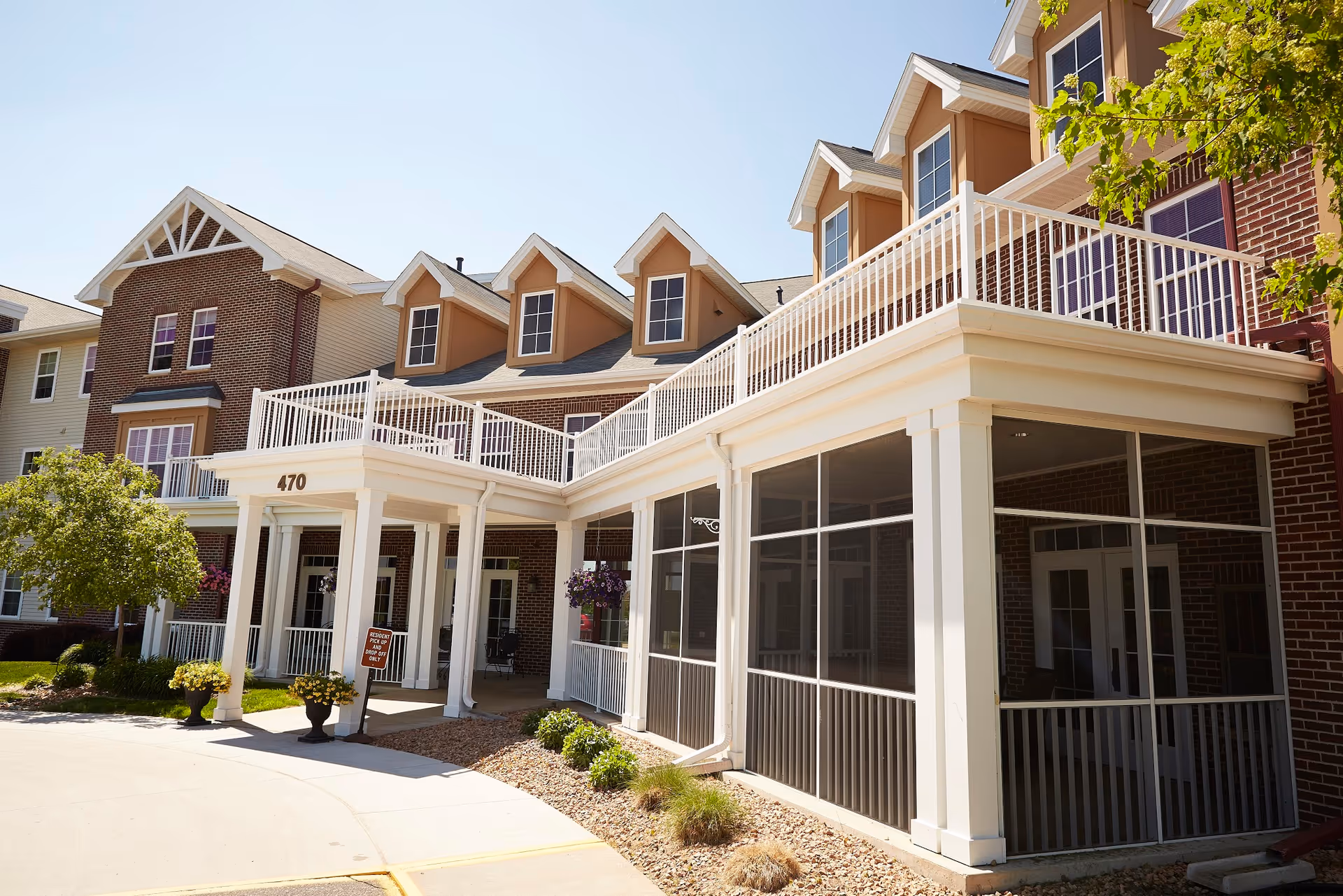 Exterior view of Deer Crest senior living facility showing a multi-story brick building with white railings, a covered entrance with the number 470, and landscaped greenery under a clear blue sky.