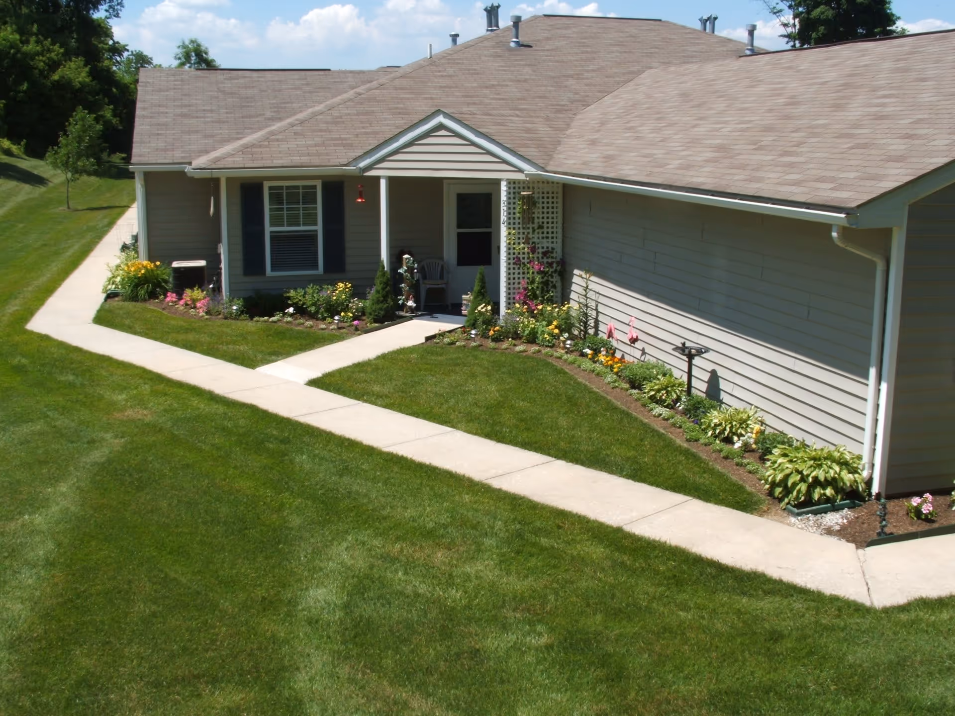Exterior view of a single-story residential building with beige siding and a brown shingled roof. The building has a small covered porch with a chair and is surrounded by well-maintained green grass and flower beds with various colorful flowers. A concrete walkway leads to the porch and extends along the side of the building. The sky is partly cloudy.