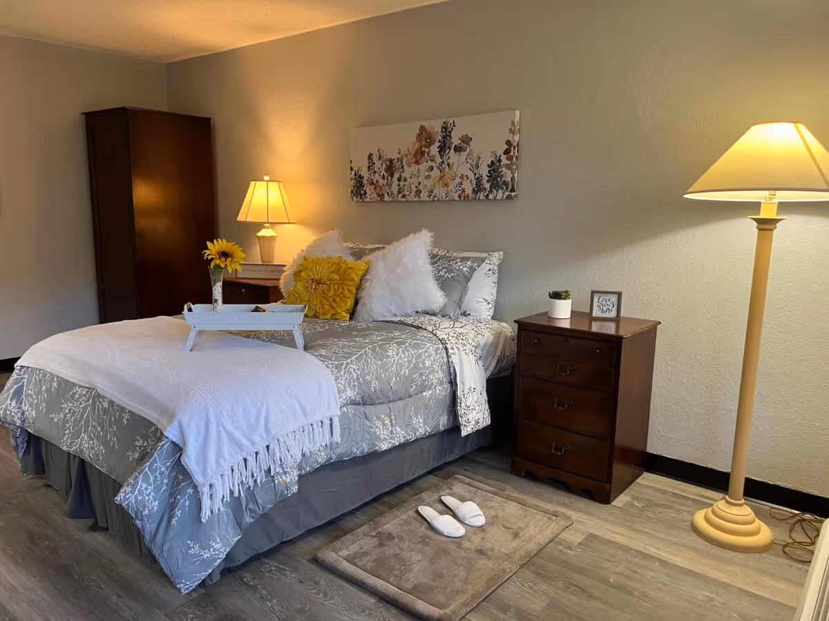 Neatly made bed with decorative pillows and a tray, flanked by a nightstand, wardrobe and two lamps in a softly lit bedroom.