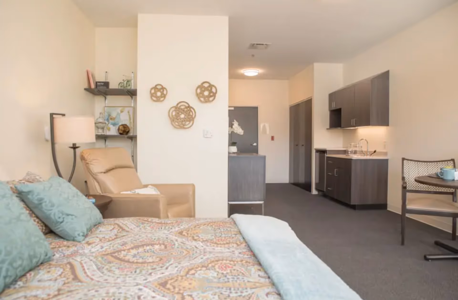A senior living facility room featuring a bed with patterned bedding and teal pillows, a beige recliner chair next to a floor lamp, a small shelving unit with decorative items, a kitchenette with dark cabinets and a sink, and a small round table with a chair and a cup on it.