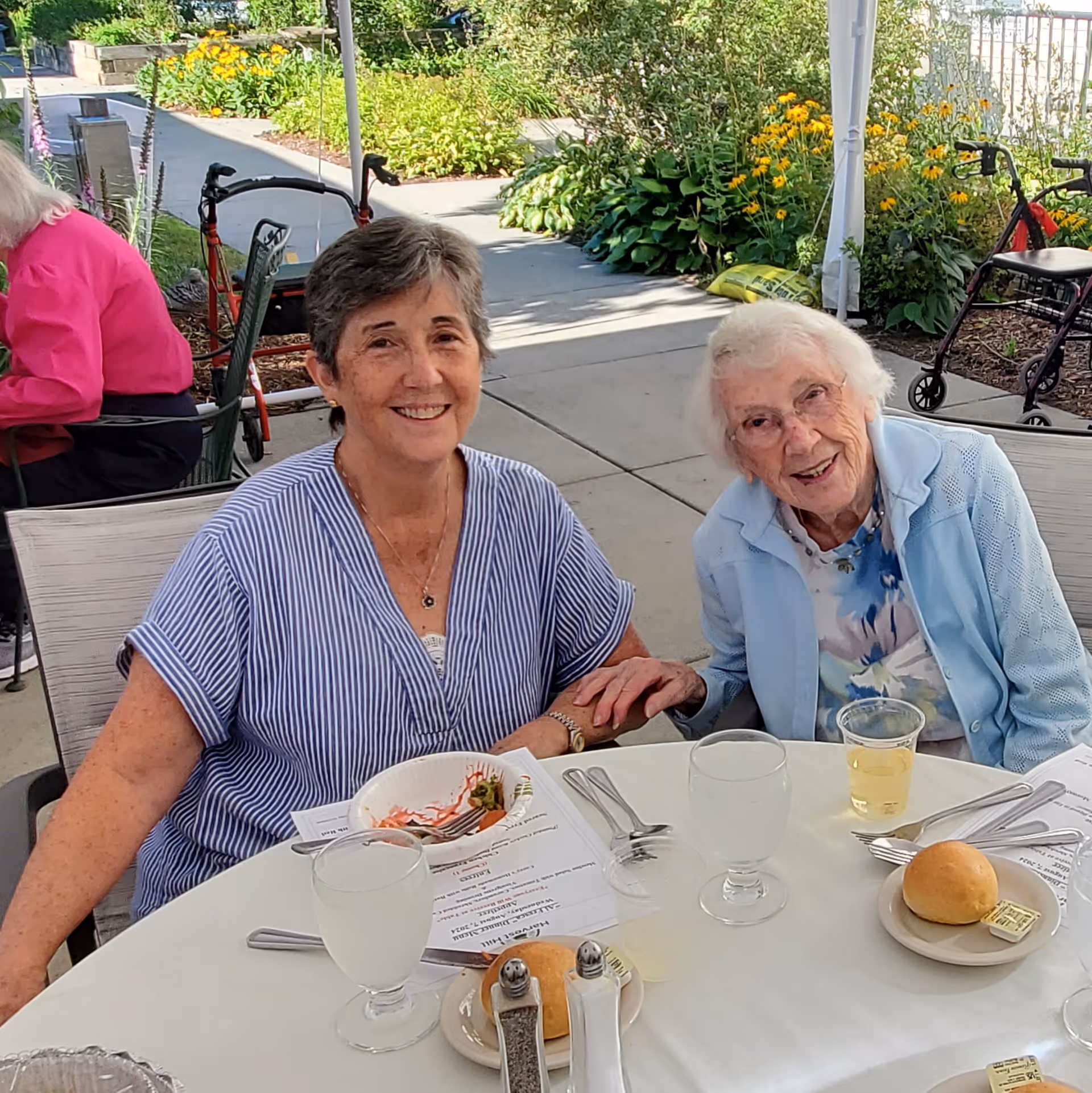 Two elderly women sitting at a round table outdoors, smiling and holding hands. The table is set with glasses of water, a bread roll with butter, utensils, and a menu. There are plants and a walkway in the background, along with mobility aids like walkers.