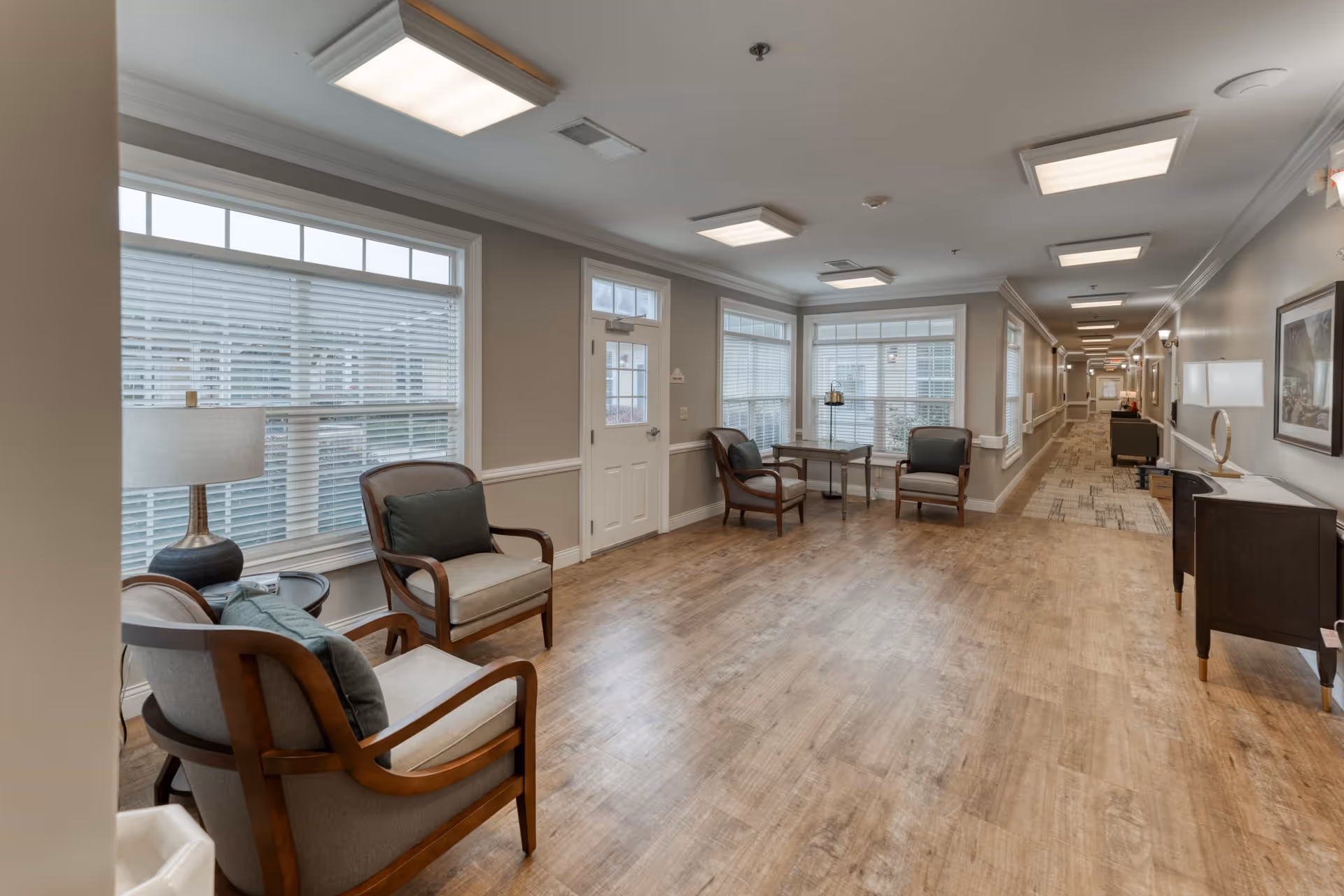 A bright and spacious hallway area in a senior living facility with wooden flooring, several armchairs with cushions, a small table, large windows with blinds, and soft overhead lighting. The hallway extends into the distance with additional seating and decor along the walls.