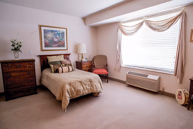 A neatly arranged bedroom with a single bed covered in beige bedding and multiple pillows. There is a wooden dresser with a vase of flowers on top to the left of the bed, a nightstand with a lamp and a small clock, and a cushioned chair with a red seat next to the nightstand. A large window with blinds and beige curtains allows natural light into the room. The carpet is light beige, and a framed floral painting hangs on the wall above the bed.