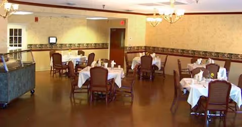 Dining room with multiple round tables covered in white tablecloths and chairs, chandeliers overhead and a buffet cart at the side.