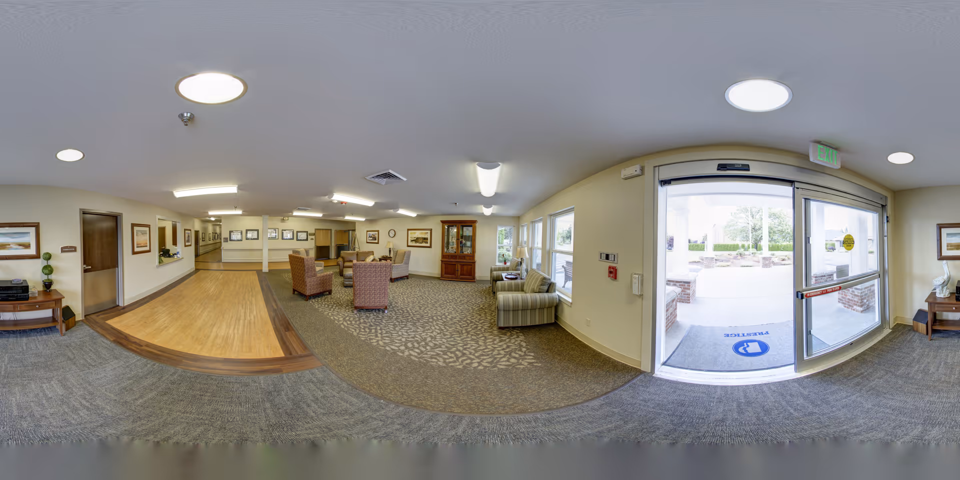 Interior view of a senior living facility lobby or common area with seating arrangements including armchairs and sofas, a wooden cabinet, framed pictures on the walls, and large windows letting in natural light. The entrance door is visible with a mat that has a logo and the word 'PRACTICE'.