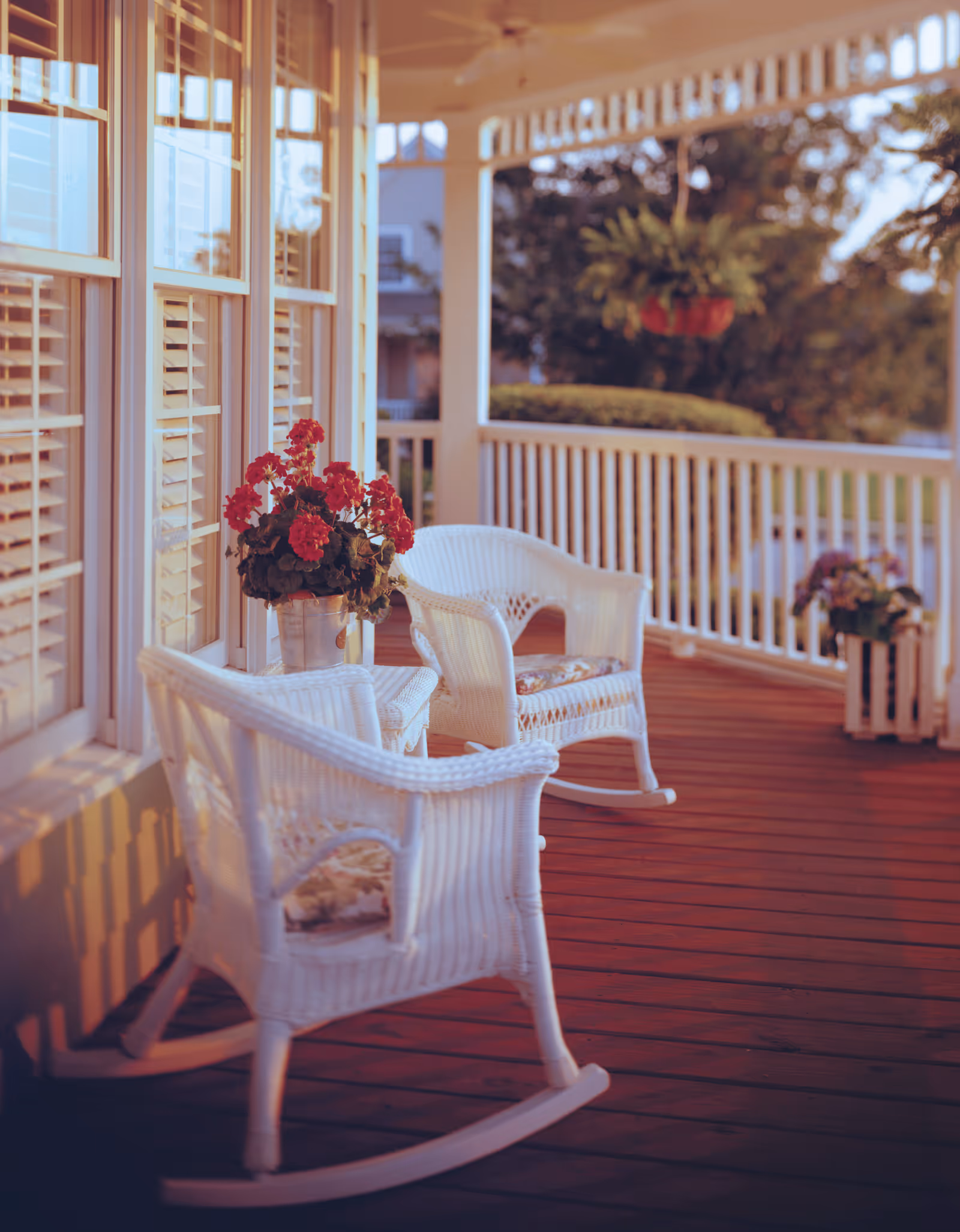 A cozy porch with two white wicker rocking chairs and a small table between them holding a pot of red flowers. The porch has wooden flooring, white railings, and windows with white shutters. There are hanging and potted plants in the background, with greenery visible beyond the porch.