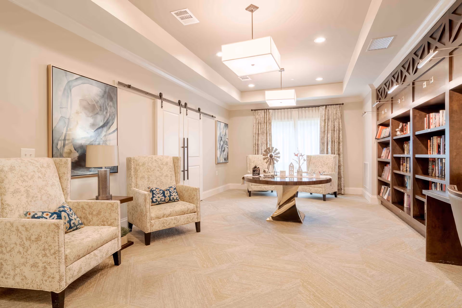 A bright and spacious living room area with beige patterned armchairs, a round wooden table with decorative items, large built-in wooden bookshelves filled with books, abstract wall art, and large windows with curtains allowing natural light to fill the room.