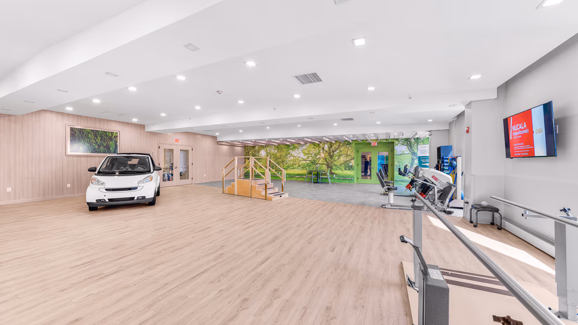 A spacious rehabilitation room with light wood flooring and white ceiling lights. On the left side, there is a white car used for therapy purposes. In the center, there is a wooden staircase with handrails for physical therapy. The back wall features a large mural of a green park with trees and a picnic table. On the right side, there are parallel bars for walking exercises and a mounted TV screen displaying information. The room is bright and designed for physical rehabilitation activities.