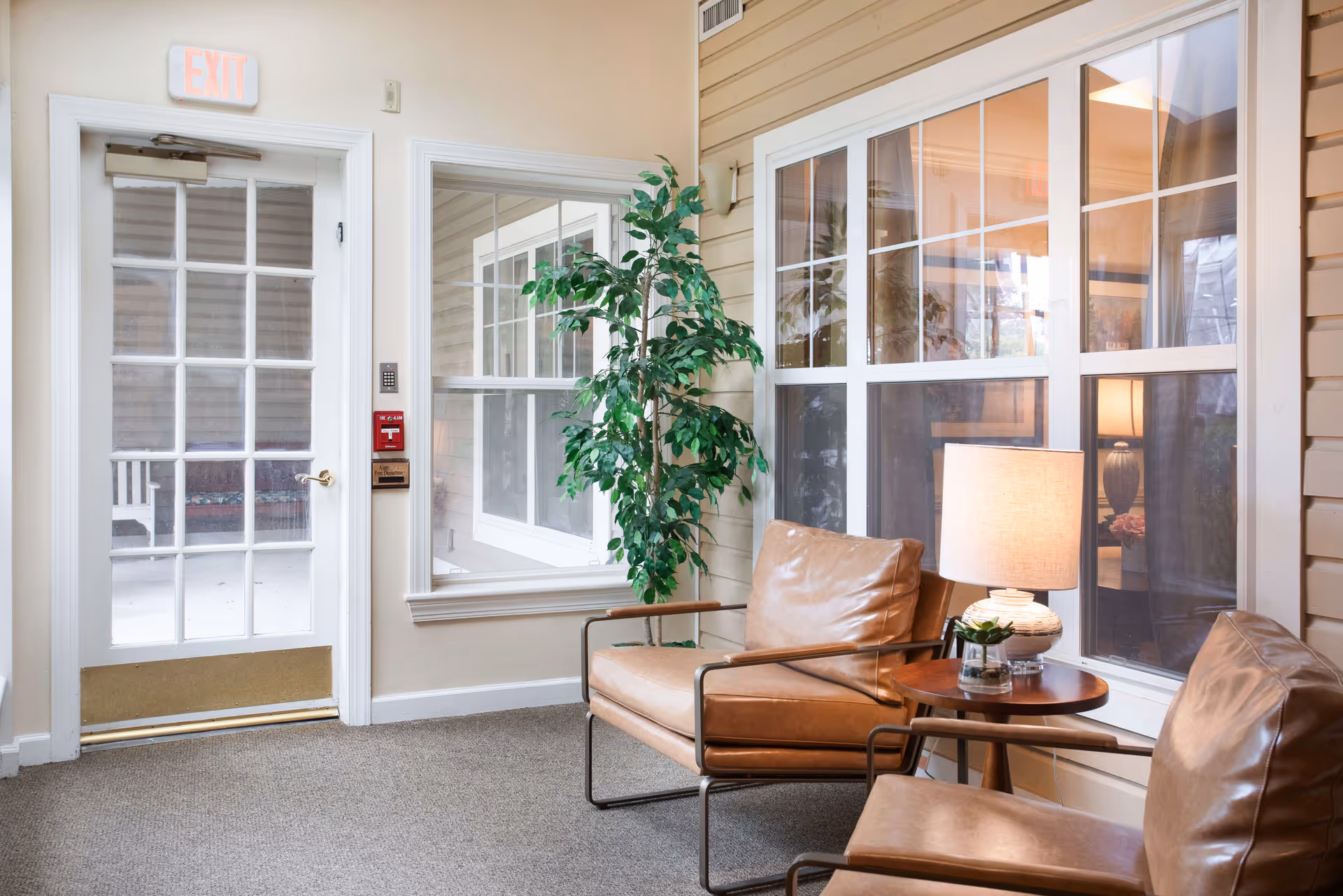 A cozy seating area with two brown leather chairs and a small round wooden table with a lamp and a small plant on it. There is a large window behind the chairs and a glass door with an exit sign above it. A tall green plant is placed near the window.