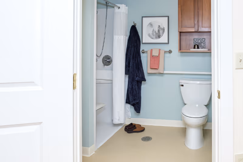 A clean and accessible bathroom with a white toilet, a shower with a white curtain, a navy blue bathrobe hanging on a hook, two towels on a towel rack, a pair of brown slippers on the floor, a framed picture on the light blue wall, and a wooden cabinet mounted above the toilet.