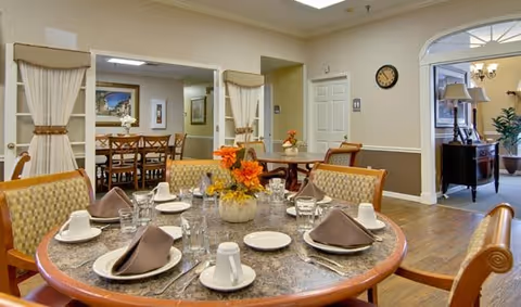 A well-lit dining room with round tables set with plates, cups, napkins, and floral centerpieces, with chairs and doorways to adjacent rooms.