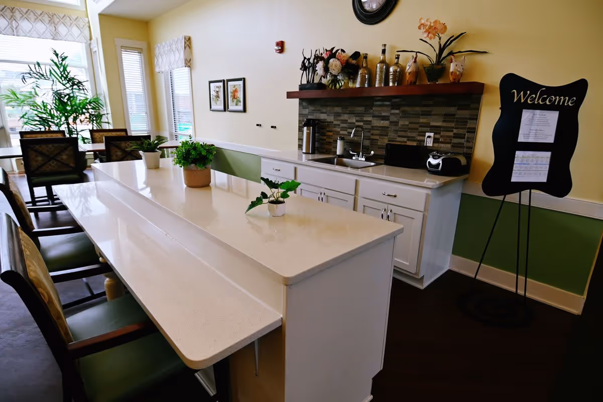 Interior view of a communal dining and kitchen area with a long white countertop island featuring three small potted plants. Behind the island is a kitchen counter with a sink, decorative bottles, flowers, and a toaster. A black welcome sign on a stand is positioned against the wall. There are chairs around the island and a dining table with chairs near large windows letting in natural light.