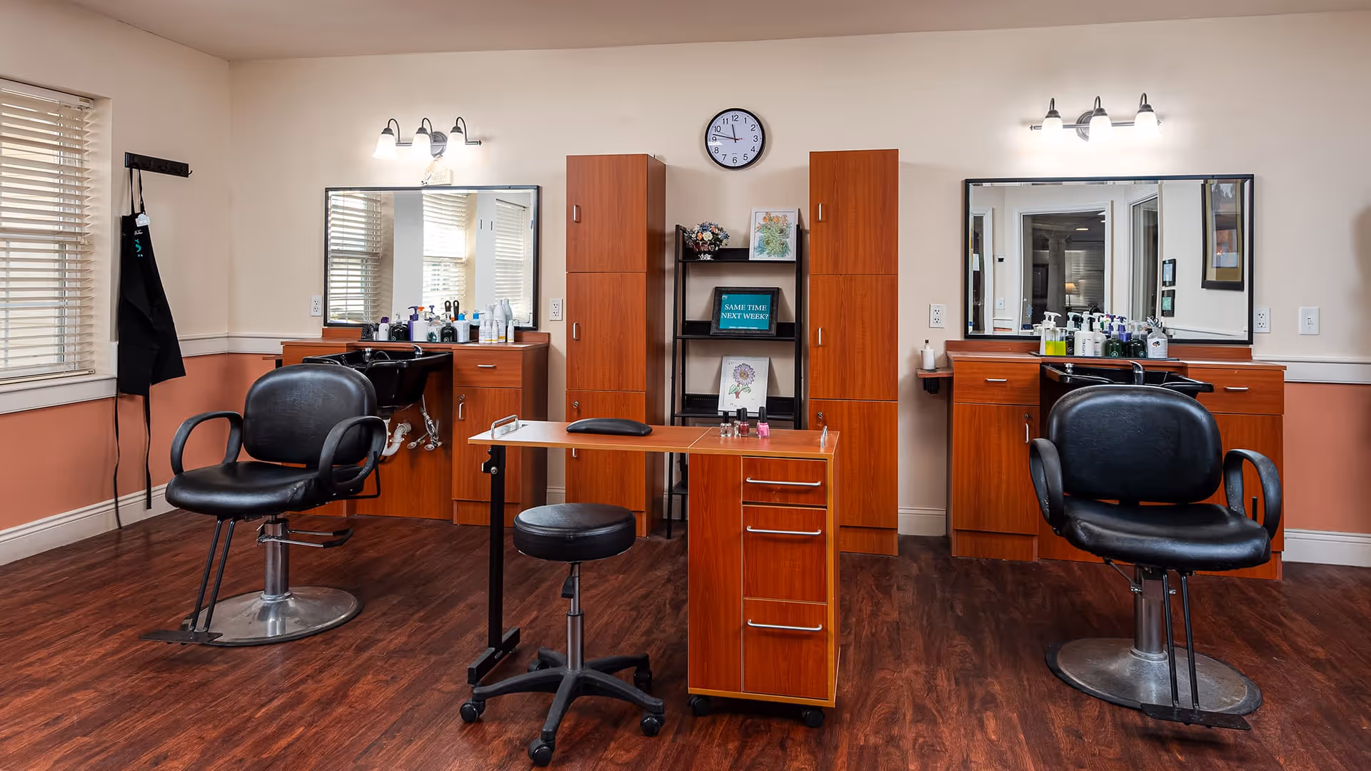Interior of a salon room with two black salon chairs in front of wooden cabinets and mirrors. There are various hair care products on the counters, a small rolling stool, and a wooden manicure table in the center. The room has wood flooring, beige and peach colored walls, and a clock above the cabinets.
