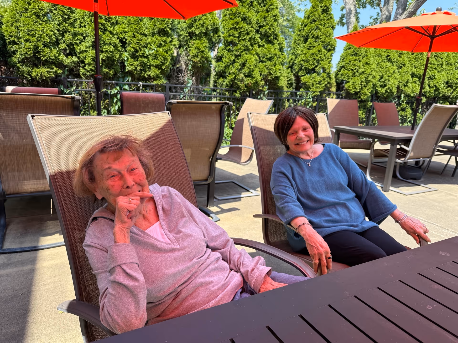 Two elderly women sitting and relaxing on outdoor patio chairs under red umbrellas, with green trees in the background on a sunny day.