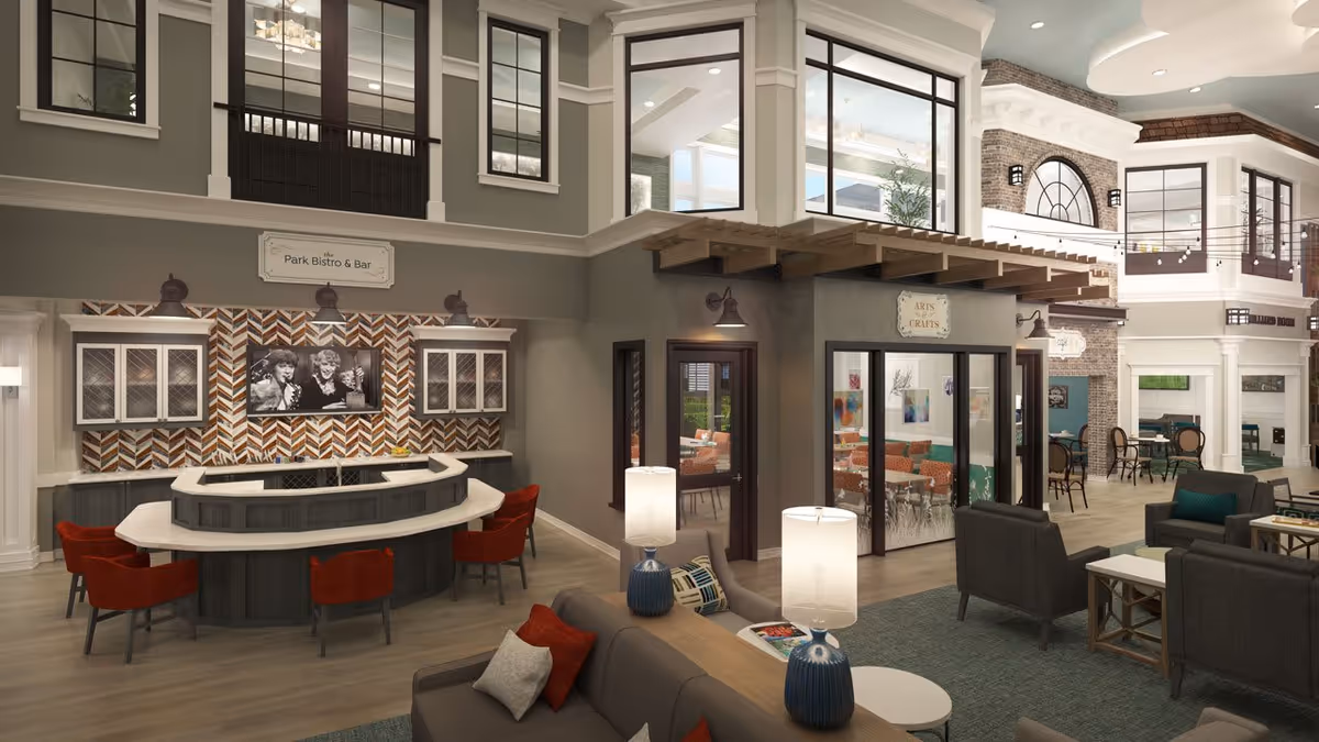 Interior view of a senior living facility featuring a cozy lounge area with sofas and armchairs, a bar area labeled 'Park Bistro & Bar' with red chairs, and a room labeled 'Arts & Crafts' with tables and chairs visible through glass doors. The space has large windows, warm lighting, and a welcoming atmosphere.