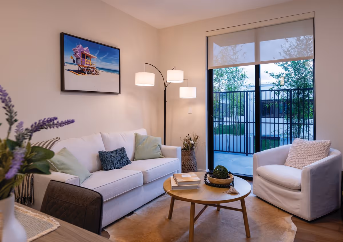 A cozy living room with a white sofa adorned with green and blue pillows, a white armchair with a textured pillow, a round wooden coffee table with books and decorative items, a floor lamp with three lights, and a large window with a view of an outdoor patio with greenery.