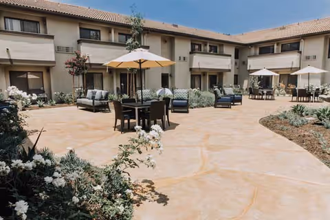 Sunlit courtyard of a senior living facility with patio tables, umbrellas, lounge seating, and a surrounding two-story building.