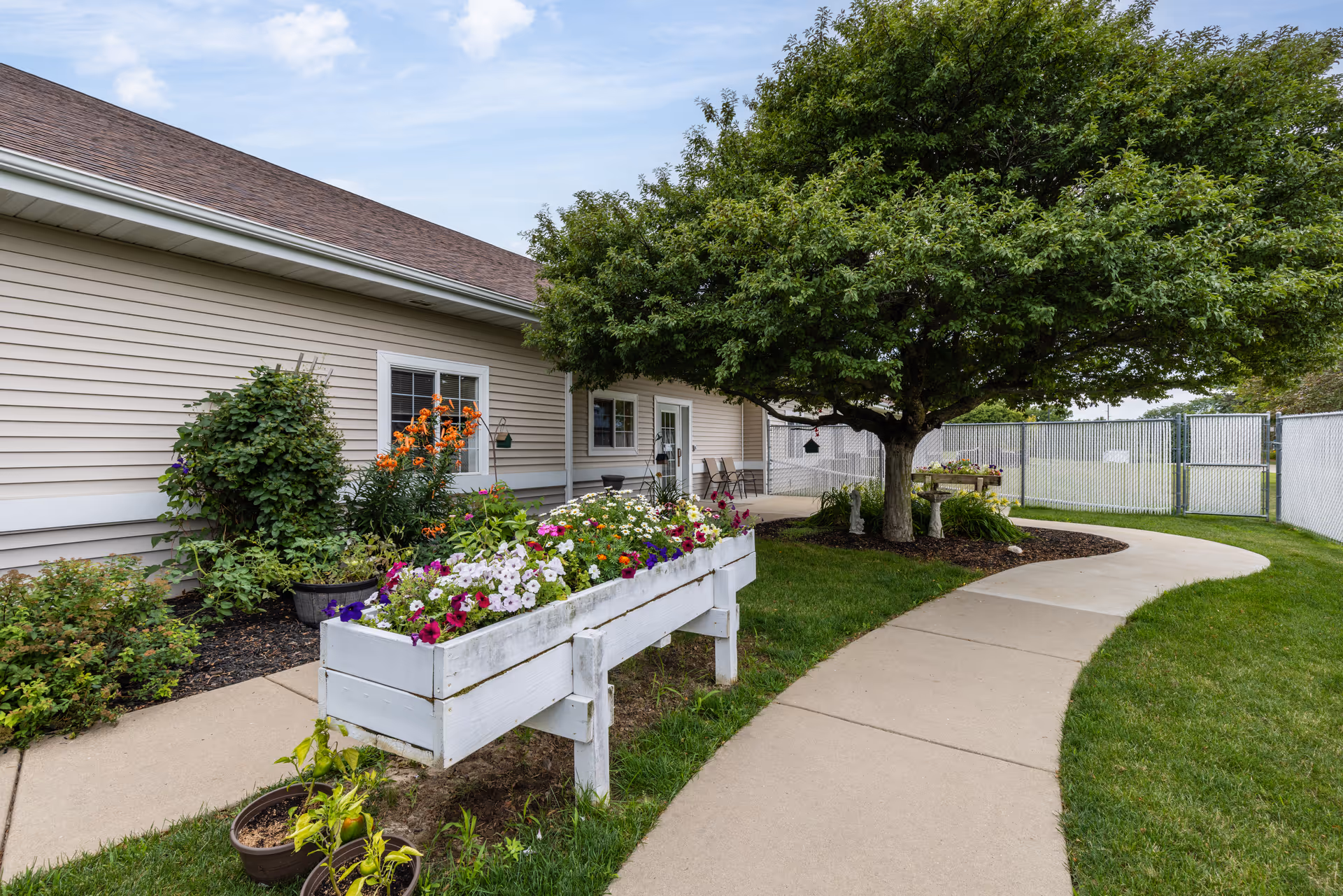 A paved walkway curves past raised flower boxes and a large tree beside a beige-siding senior living building.