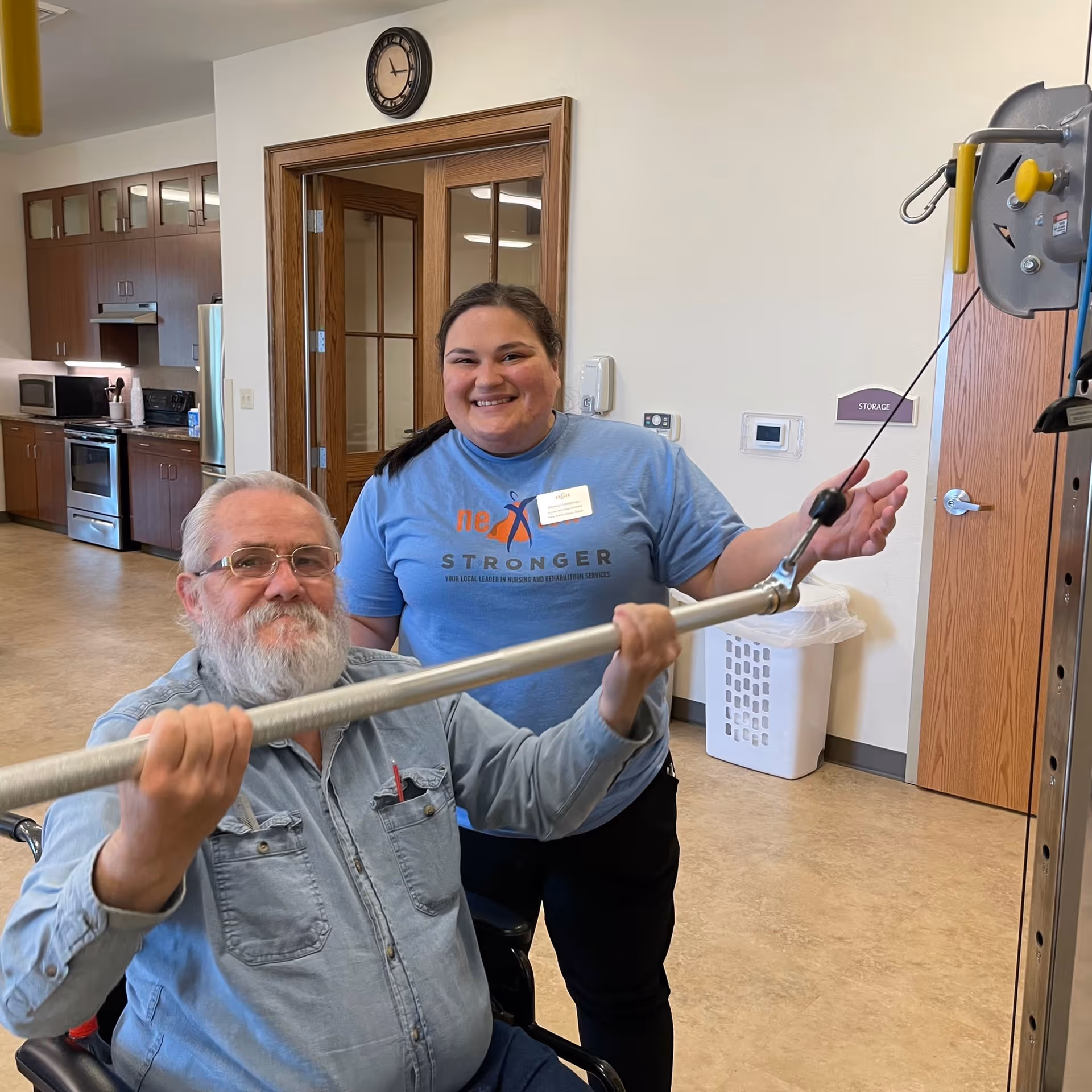 An elderly man in a wheelchair is using a lat pulldown exercise machine with assistance from a smiling female staff member wearing a blue shirt with a name tag. They are in a room with a kitchen area in the background and a clock on the wall.