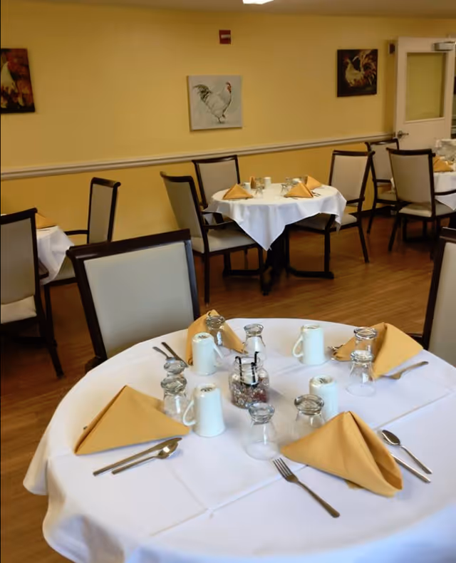 Dining room with round tables covered in white tablecloths, set with yellow folded napkins, glasses, mugs, and silverware. Chairs with light-colored cushions surround the tables. The walls are painted yellow and decorated with paintings of roosters.