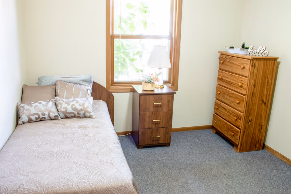 A small, simple bedroom with a single bed covered in a beige quilt and decorative pillows. Next to the bed is a wooden nightstand with three drawers, a table lamp, and a small flower arrangement. A window with wooden trim lets in natural light. Across from the bed is a wooden chest of drawers with five drawers, decorated with small plants and a decorative 'hello' sign on top. The room has light-colored walls and a gray carpeted floor.