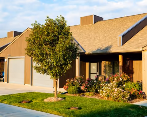 Exterior view of a senior living community building with a tree and flower bushes in front, a concrete driveway, and a clear sky above.
