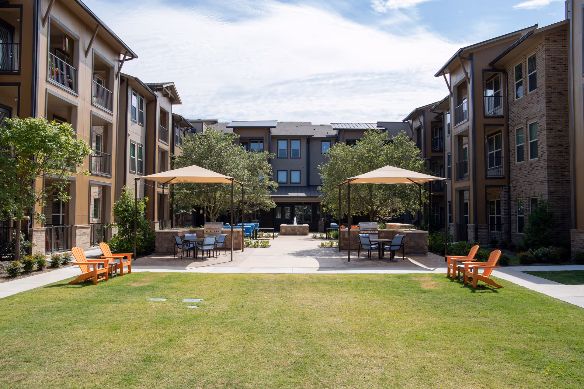 Outdoor courtyard area of a senior living facility with green lawn, two beige canopy tents with tables and chairs underneath, orange Adirondack chairs on either side, surrounded by three-story residential buildings and trees under a partly cloudy sky.