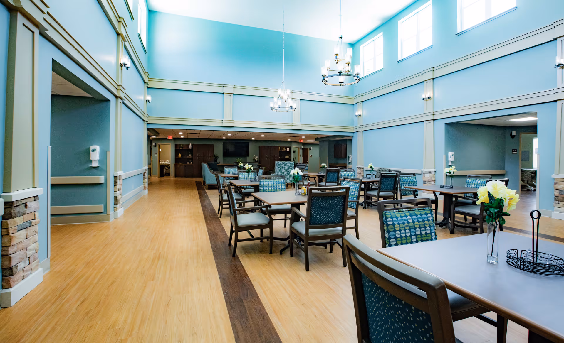 Spacious dining room with high ceilings and large windows, featuring multiple tables and chairs with patterned upholstery. The room has light blue walls, wooden flooring with a darker wood strip in the center, and chandeliers hanging from the ceiling. There are flower vases on some tables and a TV mounted on the far wall.