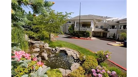 Exterior view of Hearthstone At Murrayhill facility with a landscaped garden featuring a small pond surrounded by rocks and flowering plants in the foreground, and a multi-story building with balconies and a driveway in the background.