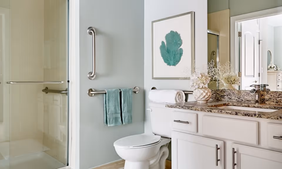 Bright bathroom featuring a glass-enclosed shower, a toilet, and a granite-top vanity with sink, teal towel and matching artwork.