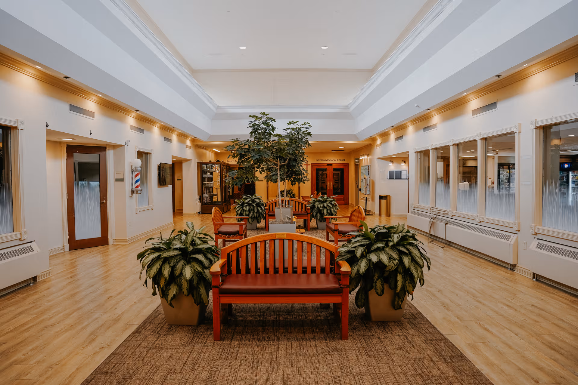 A spacious, well-lit interior common area with wooden benches and chairs arranged around large potted plants. The room has light-colored walls, a high ceiling with recessed lighting, and wood flooring. Doors and windows line the walls, and a sign above a door in the background reads 'Hickman Memorial Chapel.'