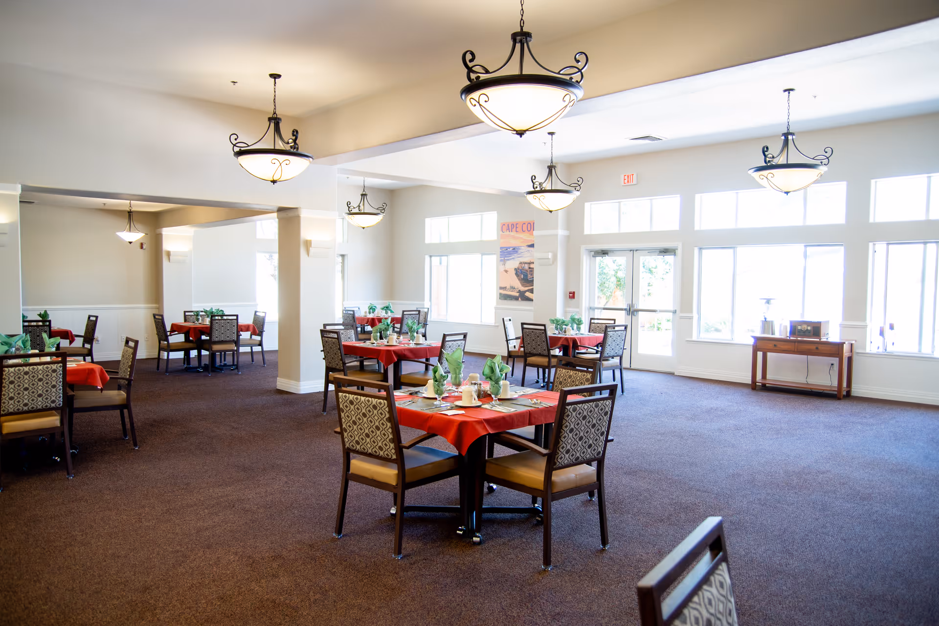 A bright and spacious dining room in a senior living facility with multiple tables covered in red tablecloths, each set with plates, cups, and green napkins. The room has large windows allowing natural light to fill the space, decorative hanging light fixtures, and carpeted floors.
