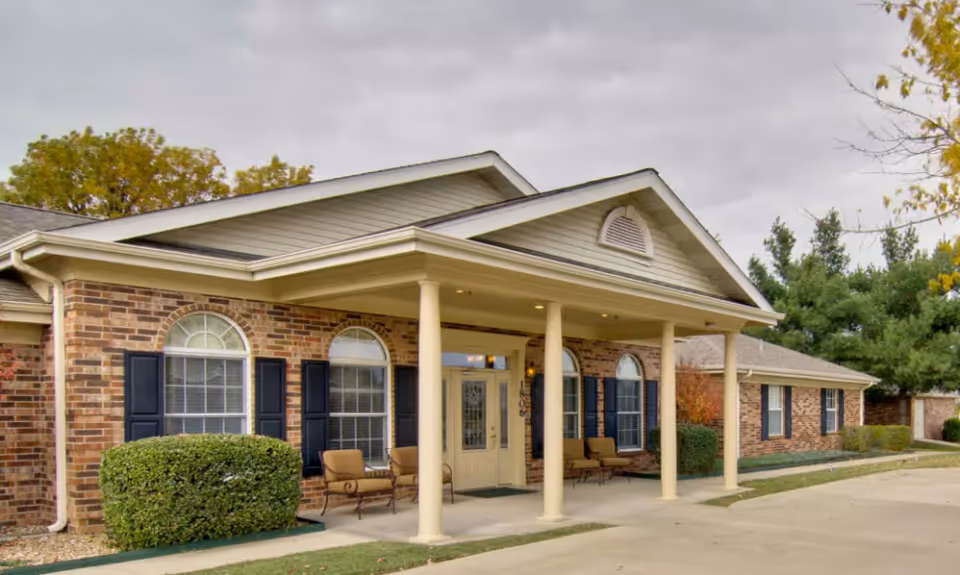 Front entrance of a single-story brick senior living facility with a covered portico, columns, seating, and arched windows.