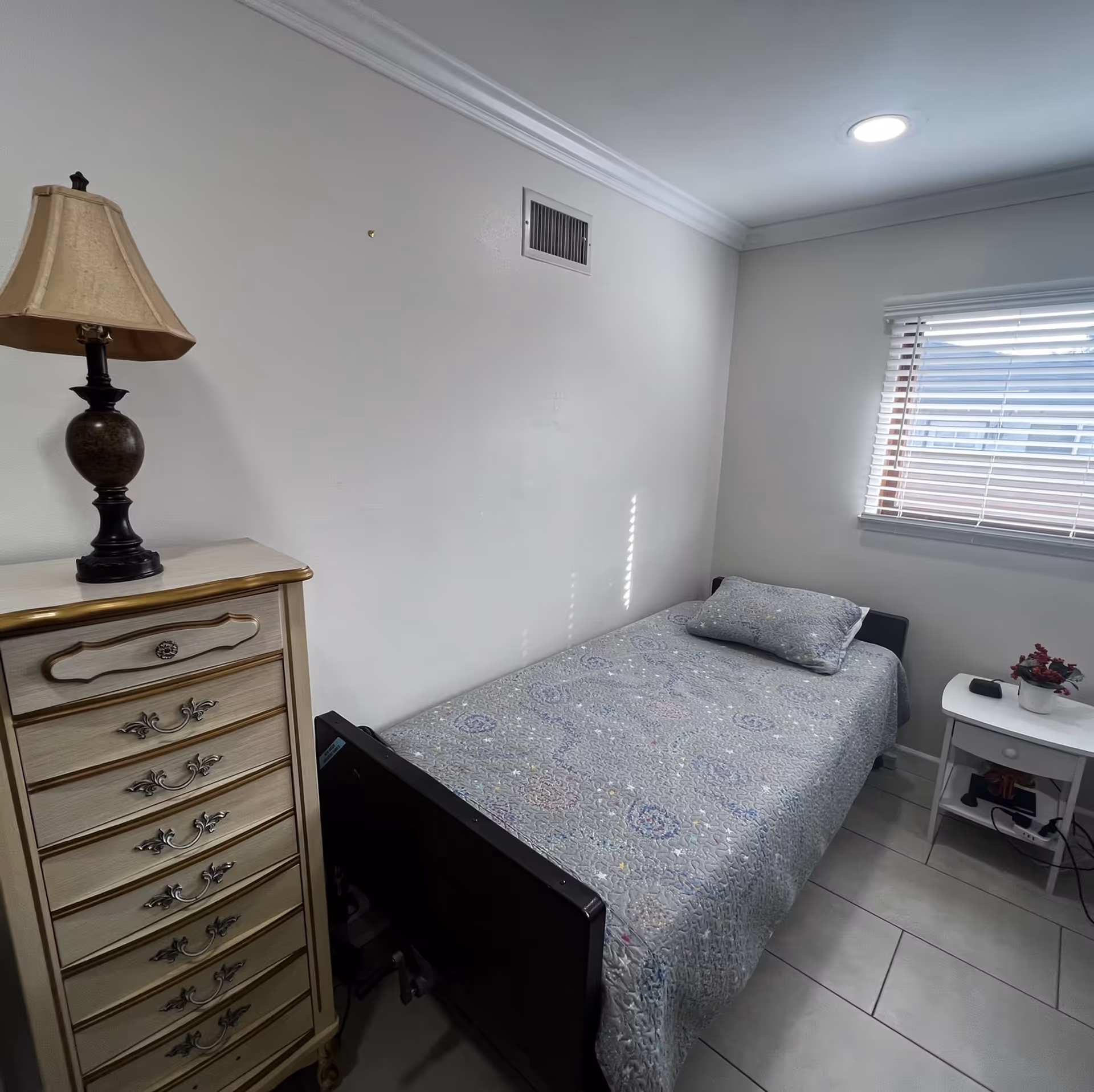A small bedroom with a single bed covered in a patterned quilt and a matching pillow. Next to the bed is a white nightstand with a small potted plant and an electronic device on it. On the left side of the image, there is a tall dresser with multiple drawers and a decorative lamp on top. A window with blinds is on the right wall, allowing natural light into the room. The walls are plain white, and the floor is tiled.