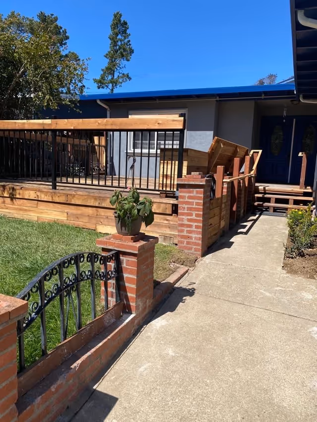 Outdoor view of a residential facility with a concrete walkway leading to a blue double-door entrance. There is a wooden deck with black metal railings and brick pillars along the walkway. A potted plant is placed on one of the brick pillars. The sky is clear and blue.
