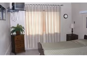 A simple bedroom with a bed covered in a beige bedspread, a wooden nightstand with a lamp, a small wooden dresser with a potted plant, a wall-mounted television, and a window with closed beige curtains.