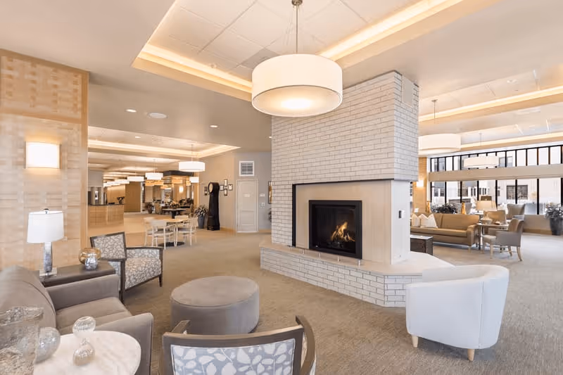 A spacious and well-lit senior living facility common area featuring a central white brick fireplace with a fire burning inside. The room is furnished with various comfortable chairs, sofas, and small tables arranged for socializing and relaxation. Large windows in the background allow natural light to fill the space, and modern ceiling lights provide additional illumination.