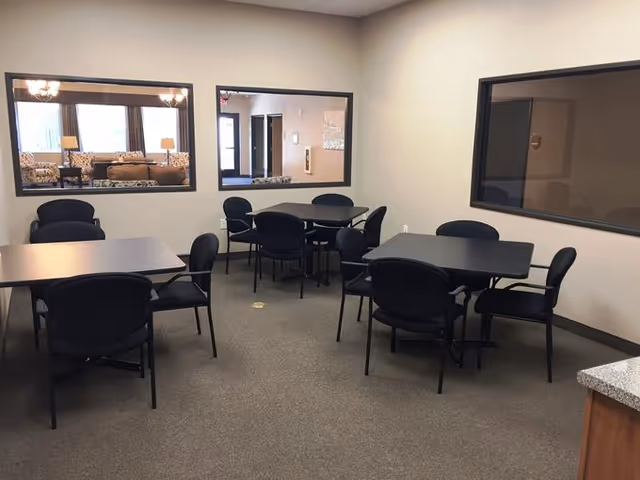 A small room with four black tables, each surrounded by black chairs. The room has beige walls and carpeted floor. There are three large windows on the walls showing adjacent rooms, one of which has sofas and lamps visible. The room is well-lit with overhead lighting.