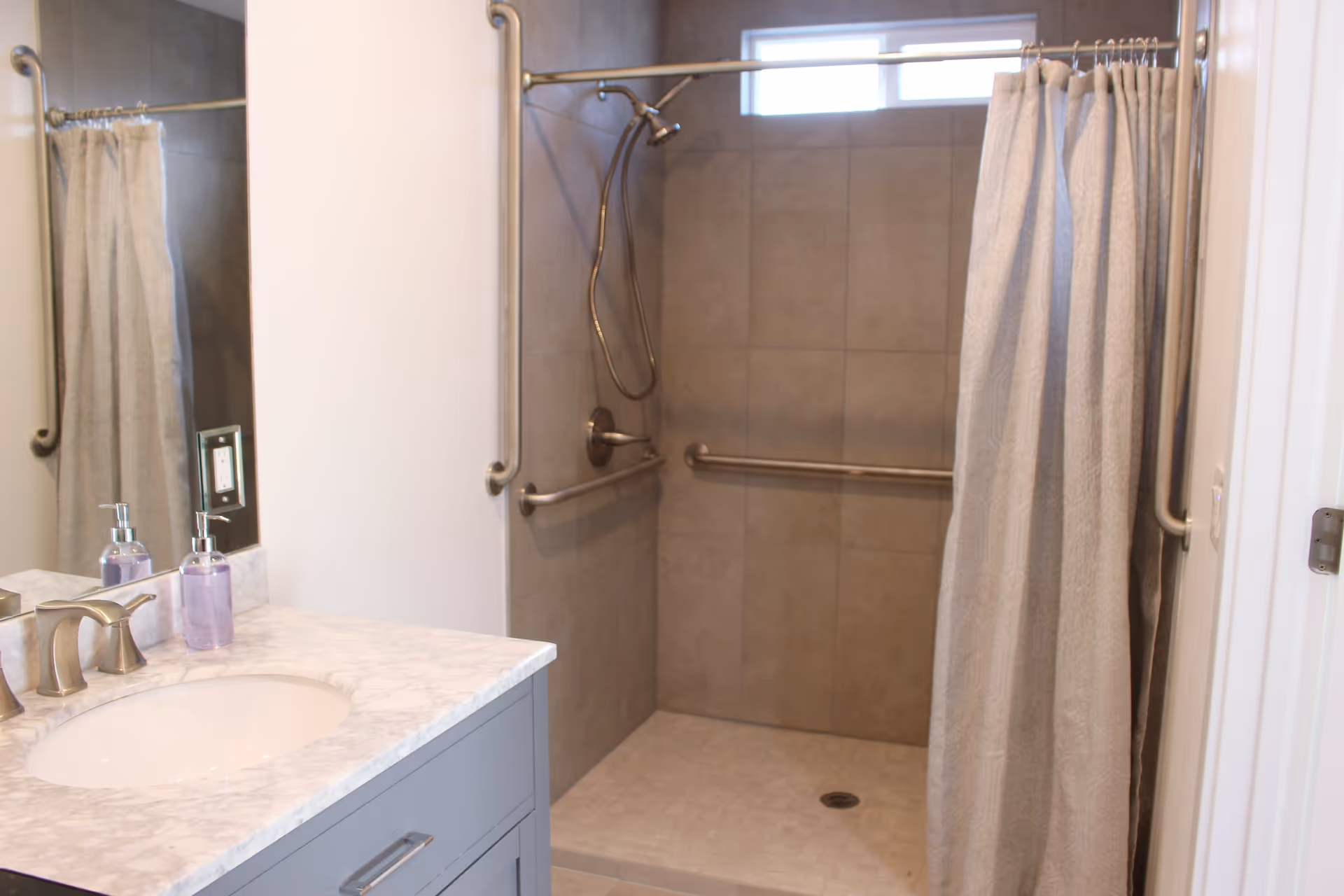 A bathroom with a marble countertop sink and a walk-in shower with beige tiles. The shower has a silver showerhead, grab bars, and a beige shower curtain. There is a small window above the shower.