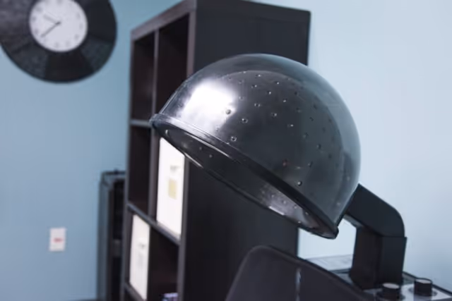 Close-up of a vintage hair dryer hood in a room with light blue walls, a black shelving unit with storage boxes, and a round wall clock.