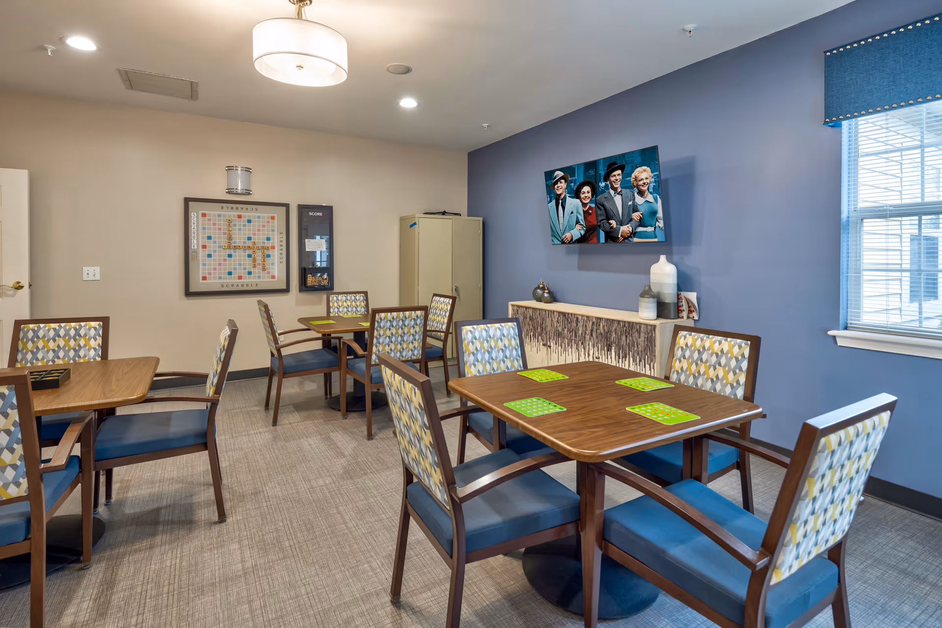 A well-lit common room with several wooden tables and chairs featuring patterned upholstery. The tables have green placemats, and a Scrabble board game is mounted on the beige wall. A blue accent wall has a mounted TV displaying a vintage black-and-white photo of four people smiling. Below the TV is a decorative sideboard with vases. A window with blinds and a blue valance allows natural light into the room.