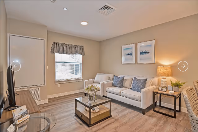 A cozy living room in a retirement community featuring a white sofa with blue and white cushions, a wooden coffee table with decorative items, a side table with a lamp and plant, a TV on a wooden stand, and two framed pictures on the beige wall. The room has a window with a valance and wood flooring.